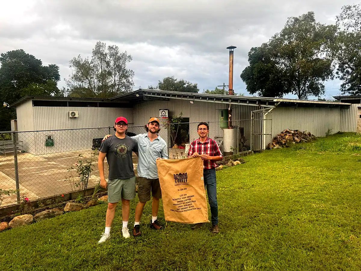 Three men outside the mill holding a burlap sack of green coffee marked direct trade, product of Honduras